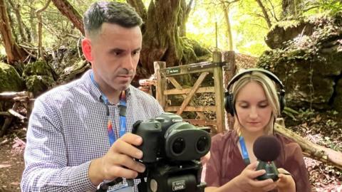 Steve with a female colleague in a woodland clearing. They are holding a camera and an audio recording device.