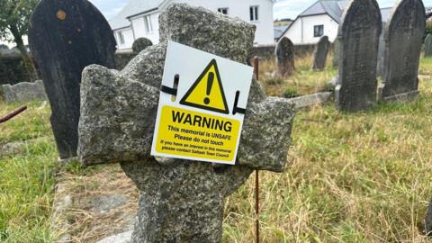 A headstone in the shape of a cross. It has a warning sign on it. It is tilted slightly to the right. The grass is green and brown. There are headstones in the background. 