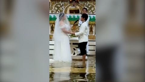 A couple in a church holding hands, wearing wedding outfits, standing in water