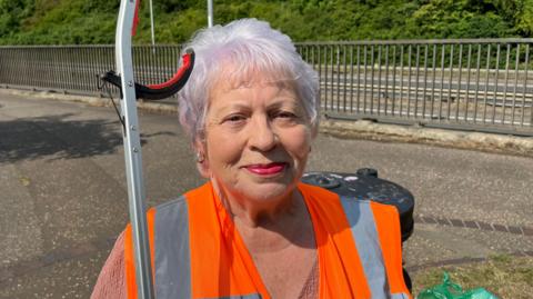 Margaret Faulkner has short grey hair with a pink tint. She has red lipstick and small gold earrings. She is wearing a high-vis orange tabard over a light pink V-neck top. She is carrying a litter grabber and the top of a rubbish bin is just behind her. Also in the background are some metal railings beside a roadway.