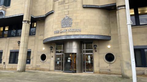 The exterior of Bradford Crown Court - a large beige stone building with steel doors at the entrance. A white flagpole is to the right of the entrance.