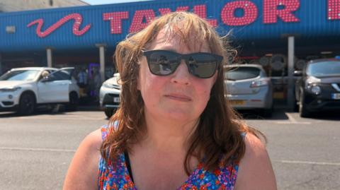 A lady in the sunshine with ginger hair, she's in a floral vest and is wearing sunglasses. She's at the sea front in Cleethorpes and she has a stern facial expression.