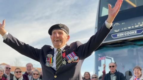 An old man wearing a military uniform and medals stands smiling with his arms outstretched in front of a bus. People are standing and smiling in the background. 