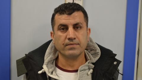 Custody image of Farhad Mohammad. He has short black hair and is wearing a grey hoody with a black coat. He is looking at the camera.