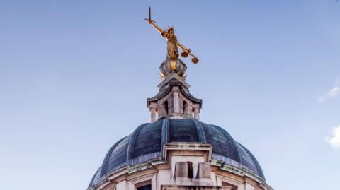 A photo of the Lady Justice statue at the top of the Old Bailey court in London. The gold statue has a sword in one hand and the scales of justice in the other hand. The top of the roof is doomed shaped and a grey colour. The other parts of the building are a cream colour.