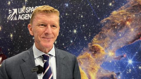 A man with light ginger hair and wearing a suit and tie stands in front of a large picture of a nebula with the UK Space Agency logo on it