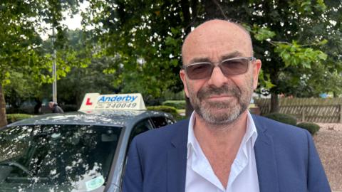 A man with tinted glasses on stood next to a blue BMW with an L plate and an advertisement board for his driving school. He has a black haired stubble with white patches and is wearing a blue blazer. The white shirt he has on has his top button un-done and he has a glum face looking at the camera. The man is stood next to his parked car in a car park. 