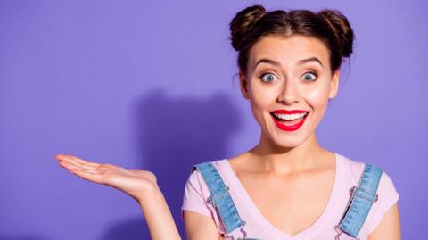 A woman smiling and holding her palm up against a lilac background.