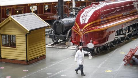 A conservator wearing a mask walks between large locomotives across a museum floor