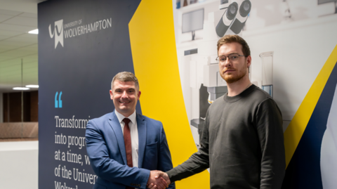 Two men shake hands while smiling at the camera. The man on the left wears a blue suit and brown tie while the man on the right wears a dark sweater.