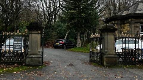 Stone pillar entrance gates to Crossley Heath School in Calderdale, with the top (finials) missing after having been stolen. The black and gold gates are open, with parked cars, grass and trees either side of the driveway.