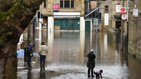 Three people and a small black dog stand on a flood-hit town centre street. A Post Office sign is visible.