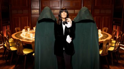 A publicity photo of presenter Claudia Winkleman, wearing a black suit and looking straight at the camera, with two hooded figures standing on either side of her. They're standing in a wood-panelled room with a circular table behind him.