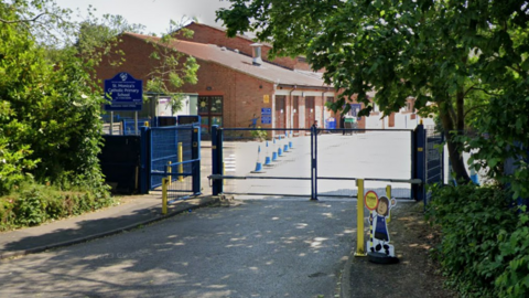 A street view image of St Monica's Catholic Primary School taken from outside its blue gates. The school building can be seen on the other side of the gates.