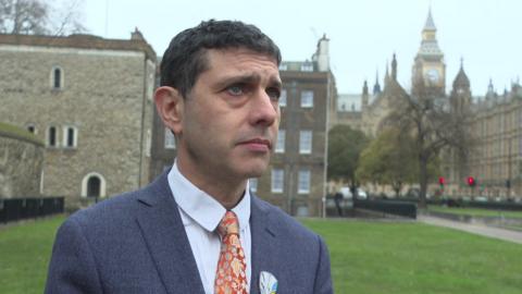 Alex Sobel stands in front of the Houses of Parliament wearing a grey suit, white shirt and floral golden tie.