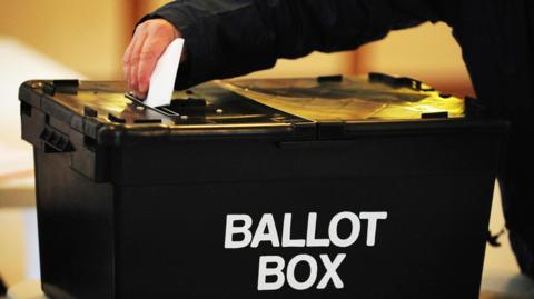 A black ballot box with a hand seen above it putting a folded sheet of paper through a slot in the top. On the side of the box the words ballot box are written in large, white capital letters.