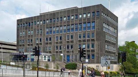 Charles Cross Police Station in Plymouth. It is a large multi-storey building near a roundabout. It is rectangular - cuboid shaped. Several people are crossing the road near the station.
