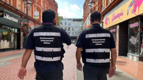 Two litter enforcement officers walk side by side with their backs to the camera. They are wearing black trousers and black shirts with a vest that has "Environmental Enforcement Officer" 