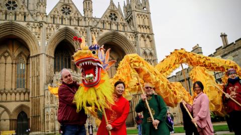 Faustina Yang in red traditional outfit, holding a golden dragon head along with four other members of the public, standing in front of the Cathedral.
