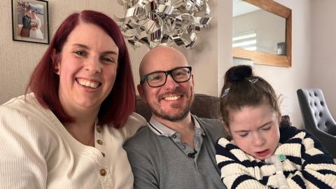 Family photo of three white people. Red haired mum Lynsey, age 44, with cream v-neck jumper sitting beside her husband Mark, age 46, who is bald with a beard and thick black glasses wearing a gray polo neck top. He is holding his daughter Thea who is wearing a black and cream striped jumper. Thea has a breathing and feeding tube coming out from her neck. They are sitting on a couch in a living room with a sliver lamp and wedding photo in the background.