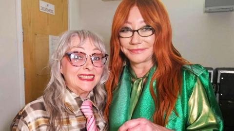 Two women standing in a dressing room at a concert. Anita Jenkins is on the left and has blonde/silver hair while wearing a patterned top in shades of beige and brown. Tori Amos is on the right - she has long red hair and a sparkly green top. She is holding a glass sculpture designed like a sweetie wrapper, with hearts in it. 