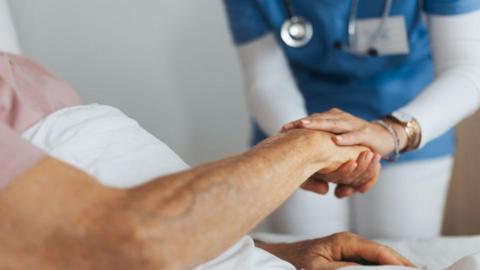 A care home residents hand being held by a care giver, wearing a blue top and white trousers. The resident is laying down in a bed. 