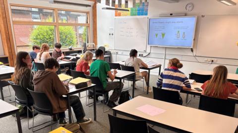 Pupils at Bedales in a classroom. There are boys and girls sat in rows of desks. They are not wearing school uniforms. There is a whiteboard at the front of the classroom. The large windows overlook a brick building and a green tree.