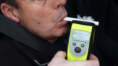 A man being breathalysed. It is a close-up shot, cropping out the top half of his face. He is blowing into a small white tube which is attached to the hand-held yellow breathalyser unit. The unit is held by someone who appears to be stood behind the camera.