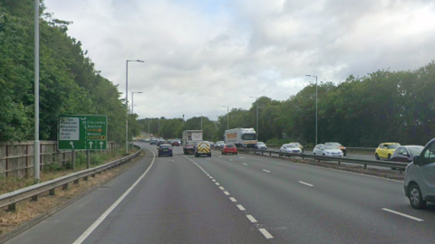 Street view of the parkway. Cars are driving on it in both directions. There is a green street sign to the left of the road showing an upcoming exit. 