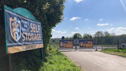The entrance to a self‑storage facility on a bright, sunny day. In the foreground on the left, a large rectangular sign is mounted on a fence and partially surrounded by greenery. The sign reads “Prince of Wales Self Storage” and features a shield‑style logo, with a phone number printed along the bottom.