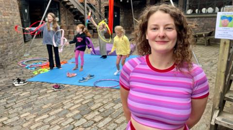 A woman in a purple striped crop top is standing in an old pottery. Behind her, mats are laid on the cobbles and four children are spinning ribbons on sticks through the air. The woman has short curly hair.