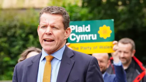 Rhun ap Iorwerth stood in front of a Plaid Cymru banner, and party activists.