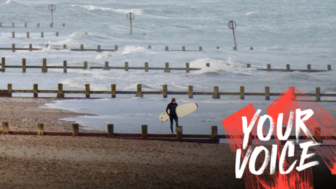 A person in a full black wetsuit carrying a surf board. They are walking along the edge of the water at Aberdeen beach, where there are large waves.