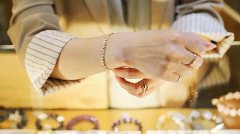 A woman with a striped brown and white shirt and pale blazer tries on a gold bracelet, with other bracelets on the table in front of her. 