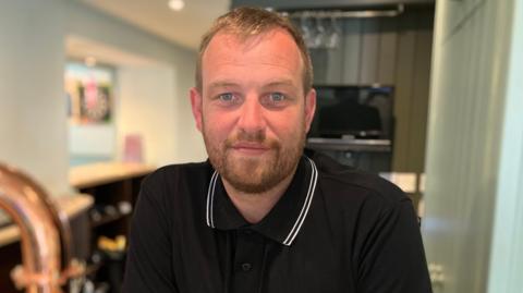 Landlord Lewis Challen stood behind his bar. He is wearing a black polo shirt which has a white collar. He is smiling and looking straight at the camera.