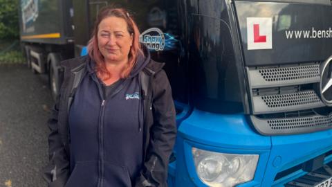 Andrea stands in front of a truck and smiles at the camera. She has shoulder length pale red hair, wears a navy blue jumper and a black jacket.