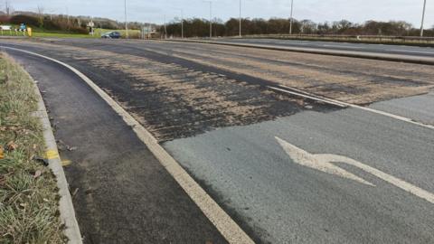 A road with two lanes. The left lane had a white arrow pointed left painted on it. The road looks worn down. There is a grass verge on the left of the road and there is a roundabout in the distance.