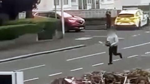 Grainy footage capturing a man wearing black trousers and a grey top running down a residential street while brandishing a chainsaw. A parked police car and a lone officer can be seen in the background