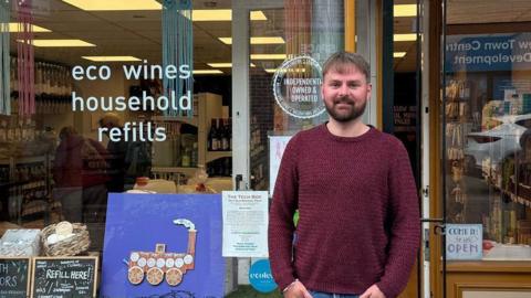 James Baggaley is stnading , smiling in front of his shop, Earth Warriors. He has short brown hair and a beard, and is wearing a dark red jumper. A sign on the window reads: "eco wines, household refills".