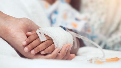 A child's hand with a medical lead in, with a bandage, on a hospital bed. The child's hand is being held by an adult. 