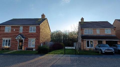 91ȱs on Silverdale Gardens. Two detached houses stand on either saide of a patch of grassland. A row of trees can be seen in the gap running behind the houses. The street sign stands by the pavement.