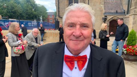 A man with white hair is standing outside a church. He is looking into the camera. He is wearing a dark suit, white shirt and red bow tie.