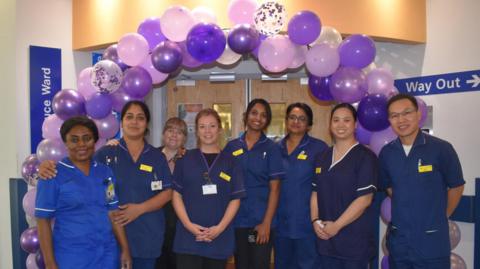 A group of staff members wearing scrubs stand beneath a purple balloon arch and smile at the camera. 