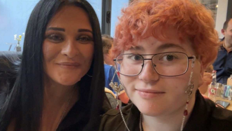 Two women side by side, looking at the camera and smiling. The woman on the right has short dyed-pink hair, glasses and is wearing a black top. The woman on the left has long dark brown hair and is wearing a black top. Behind them are tables with other people sat at them.