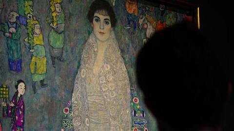 A man stands in front of a full-length Art Nouveau portrait of a woman.