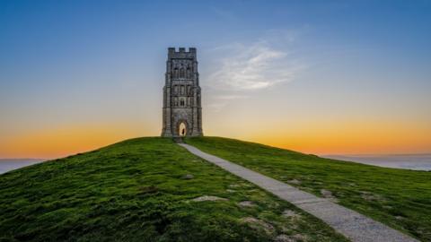 Glastonbury Tor at sunrise. The tower is on top of a hill with a path leading up to it. 