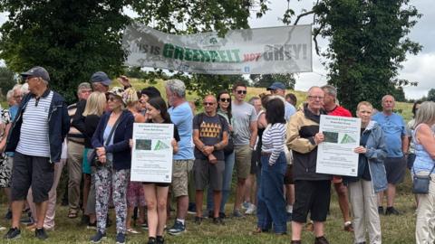 A gathering of people next to the land in Kingswinford known as The Triangle. Some people are holding signs opposing the development plans, with a banner in the background saying "this is green belt not grey".