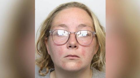 Police custody image of Stacey Sharples who has shoulder length blond hair and pale pink rimmed glasses and wearing a grey t-shirt.