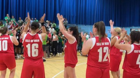 Six female basketball players in red kit face a crowd in a sports hall clapping their hands above their heads. The back of the players' vests have different numbers and the words Isle of Man printed on them in white writing.