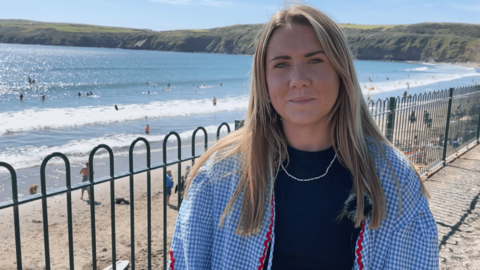 Mirain Llŷn Roberts standing near the seafront in Aberdaron, and behind her people are on the beach and in the water